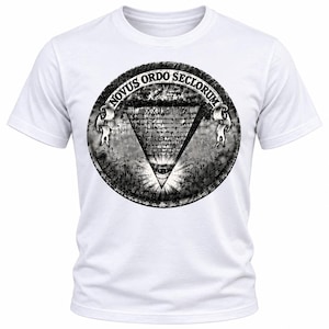 Puede incluir: Una camiseta blanca de manga corta con un gran gráfico circular en blanco y negro en el pecho. El diseño muestra una pirámide con un ojo en la base, rodeada de patrones intrincados y una cinta con el texto NOVUS ORDO SECLORUM.