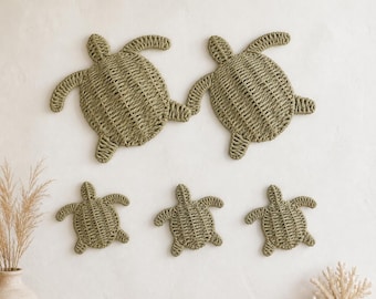 May include: A set of five wall-mounted sea turtle decorations crafted from woven sage green fiber. The collection includes two larger turtles positioned above three smaller turtles, all featuring a detailed, textured, braided pattern.