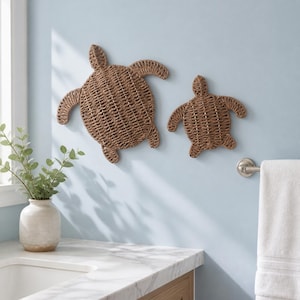 May include: Two wall-mounted sea turtle decorations crafted from woven brown natural fibers. The larger turtle is positioned to the left of the smaller turtle, both featuring detailed shell patterns and extended flippers in a flat design.
