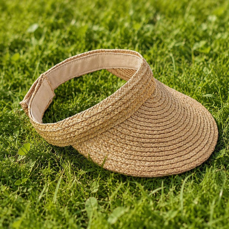 Adjustable raffia straw sun visor with velcro closure lying on green grass, wide brim view