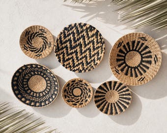 Puede incluir: Una colección de seis cestas redondas tejidas de varios tamaños, con patrones geométricos en color beige natural y negro. Las cestas muestran diversos diseños que incluyen rayas radiales, zigzags y motivos en espiral de fibra natural.
