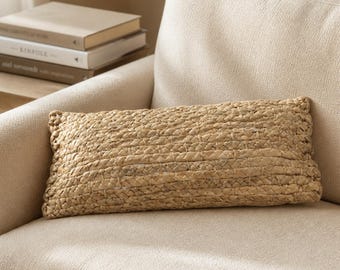 May include: A rectangular throw pillow featuring a woven tan natural fiber exterior with a braided texture. The pillow has a structured, elongated shape and a consistent light brown color throughout its surface, displaying a detailed interlaced pattern.
