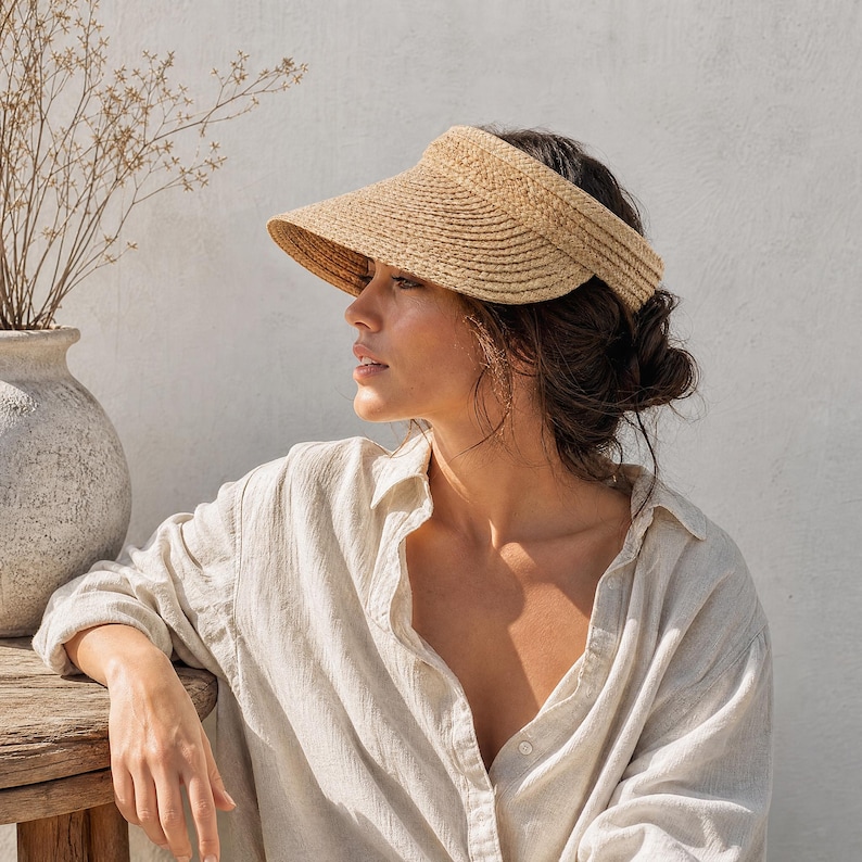 Adjustable raffia straw sun visor with wide brim worn by a woman in a white shirt on a wooden bench