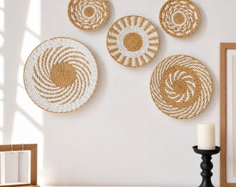 Puede incluir: Conjunto de cinco cestas de pared tejidas de diferentes tamaños. Las cestas presentan patrones espirales y solares naturales y blancos. La disposición se muestra en una pared blanca, creando una decoración mural texturizada.