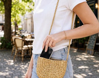 May include: A semi-circular woven straw crossbody bag with a braided strap. The bag features a natural tan color and a textured, radial pattern. A black smartphone is partially inserted into the open top of the bag, which is worn over a white t-shirt.