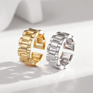Puede incluir: Dos anillos ajustables con un diseño de correa de reloj. Un anillo es dorado y el otro es plateado.