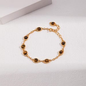 May include: A gold-toned chain bracelet featuring multiple eye-shaped links, each set with a round, brown, chatoyant gemstone. The bracelet includes a delicate link chain, a lobster claw clasp, and an adjustable extender chain with a small charm.