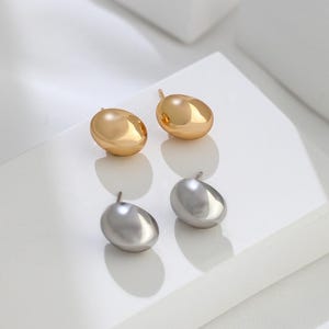 May include: A pair of earrings with a simple, modern design. One pair is gold and the other is silver. Each earring is a smooth, oval-shaped stud.