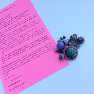 May include: A bright pink instructional sheet titled How To Properly Glaze Your Paper Bead with black text, accompanied by a collection of multicolored, multi-shaped paper beads in various sizes, including a large blue spherical bead.