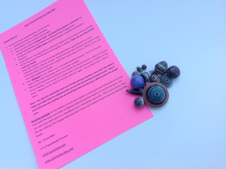 May include: A bright pink instructional sheet titled How To Properly Glaze Your Paper Bead with black text, accompanied by a collection of multicolored, multi-shaped paper beads in various sizes, including a large blue spherical bead.