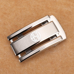 May include: A rectangular silver-toned metal belt buckle with a brushed finish and polished edges. The center features a circular engraved monogram with the letters BSD. The buckle has a mechanical sliding mechanism visible on the sides.