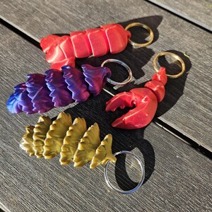 Puede incluir: Cuatro llaveros diferentes, cada uno con un diseño diferente. El primer llavero es un langostino rojo, el segundo es un langostino morado y azul, el tercero es una piña dorada y el cuarto es un langostino rojo. Todos los llaveros tienen anillas de llavero plateadas.
