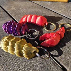 Puede incluir: Cuatro llaveros con diferentes diseños. Uno es un dragón morado y rosa, otro es un dragón dorado y los otros dos son garras de langosta rojas.