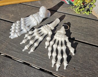 May include: Three white plastic skeleton hand and foot keychains. The foot keychain is a full foot, while the hands are just the hand bones. The keychains are on a wooden surface.