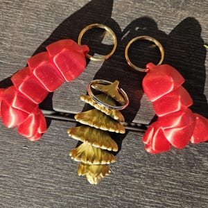 Puede incluir: Tres llaveros: dos llaveros rojos en forma de langosta y un llavero dorado en forma de árbol. Los llaveros de langosta están hechos de un material liso y brillante y tienen un cuerpo segmentado. El llavero de árbol está hecho de un material texturizado y metálico y tiene un diseño en capas.