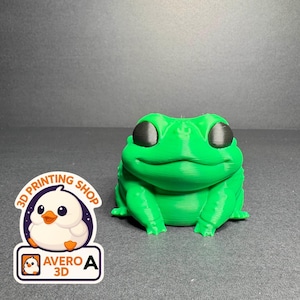 Könnte beinhalten: Eine hellgrüne, 3D-gedruckte Froschfigur mit großen, schwarzen Augen und einem lächelnden Gesichtsausdruck. Der Frosch sitzt auf einer dunklen Oberfläche. Ein Aufkleber mit einer Cartoon-Ente und dem Text "3D PRINTING SHOP" und "AVERO 3D" ist ebenfalls im Bild.