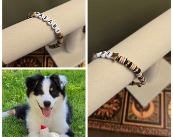 Könnte beinhalten: Ein weißes Perlenarmband mit schwarzen Buchstaben, die "WASABI" buchstabieren, und einem goldenen Sternanhänger. Das Armband wird auf einer weißen Oberfläche präsentiert.