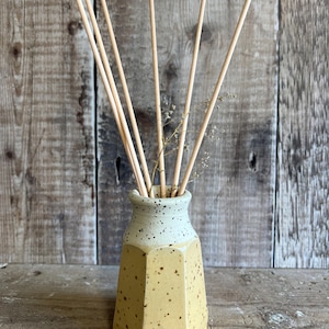 May include: A yellow and cream ceramic diffuser vase with a speckled design. The vase has a geometric shape and contains wooden diffuser sticks and dried flowers. The background is a wooden surface.