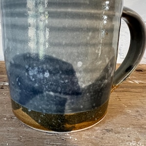 May include: A handcrafted ceramic mug with a distinctive layered design. The mug showcases a dark blue and grey colour scheme with a brown base and handle. Ideal for coffee, tea, or any hot drink.