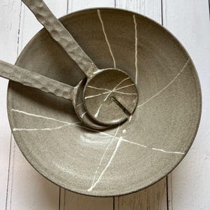 May include: A gray ceramic serving bowl with two matching serving spoons. The bowl and spoons have a textured surface and white linear designs. The spoons are resting inside the bowl.