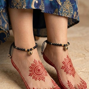 May include: A pair of black beaded anklets featuring small gold-toned metallic spacers and a central bell charm. The anklets are secured with black cords and are worn on feet decorated with intricate red circular henna patterns and a gold toe ring.