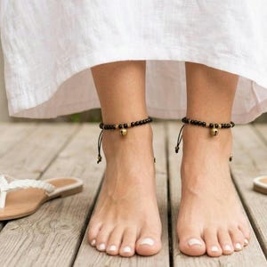 May include: A pair of black beaded anklets featuring small metallic gold-toned bells. Each anklet is constructed with a series of uniform black beads and accented with gold-colored spacers, secured with a thin black cord tie at the back.