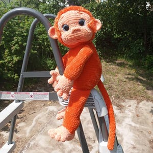May include: A handmade orange crochet monkey doll with a long tail, sitting on a metal ladder. The monkey has large, blue eyes and is made of soft, textured yarn. The doll's hands and feet are a lighter shade of orange.