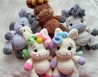 May include: A collection of handmade crochet unicorn plushies in various colors. The unicorns have soft, textured bodies, with details like bows, manes, and hooves in shades of blue, pink, purple, and brown. They are arranged on a fluffy white surface.