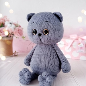 May include: A gray knitted plush cat toy with large eyes and a black nose. The cat is sitting with its legs crossed and has a soft, cuddly appearance.