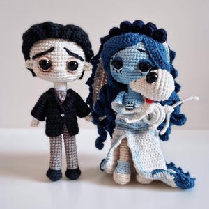 Puede incluir: Dos mu&ntilde;ecos de ganchillo hechos a mano, inspirados en una pel&iacute;cula. El mu&ntilde;eco masculino lleva un traje negro y tiene el pelo negro. La mu&ntilde;eca femenina es azul con un vestido blanco y sostiene un esqueleto blanco. Los mu&ntilde;ecos est&aacute;n sobre una superficie blanca.