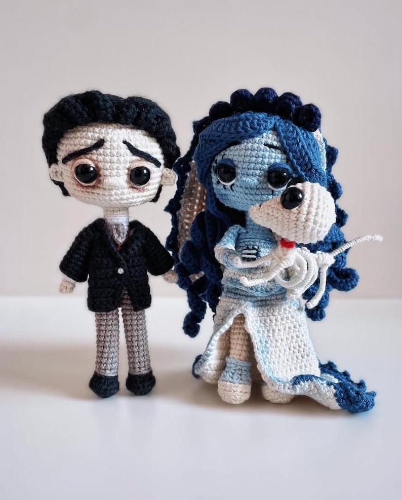 Puede incluir: Dos mu&ntilde;ecos de ganchillo hechos a mano, inspirados en una pel&iacute;cula. El mu&ntilde;eco masculino lleva un traje negro y tiene el pelo negro. La mu&ntilde;eca femenina es azul con un vestido blanco y sostiene un esqueleto blanco. Los mu&ntilde;ecos est&aacute;n sobre una superficie blanca.