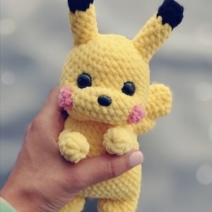 Może przedstawiać: Ręcznie robiona żółta pluszowa zabawka Pikachu z czarnymi uszami i oczami oraz różowymi policzkami. Pluszak trzymany jest w dłoni, prezentując swój miękki, teksturowany wygląd. Uroczy i przytulny przedmiot o tematyce Pokemon.