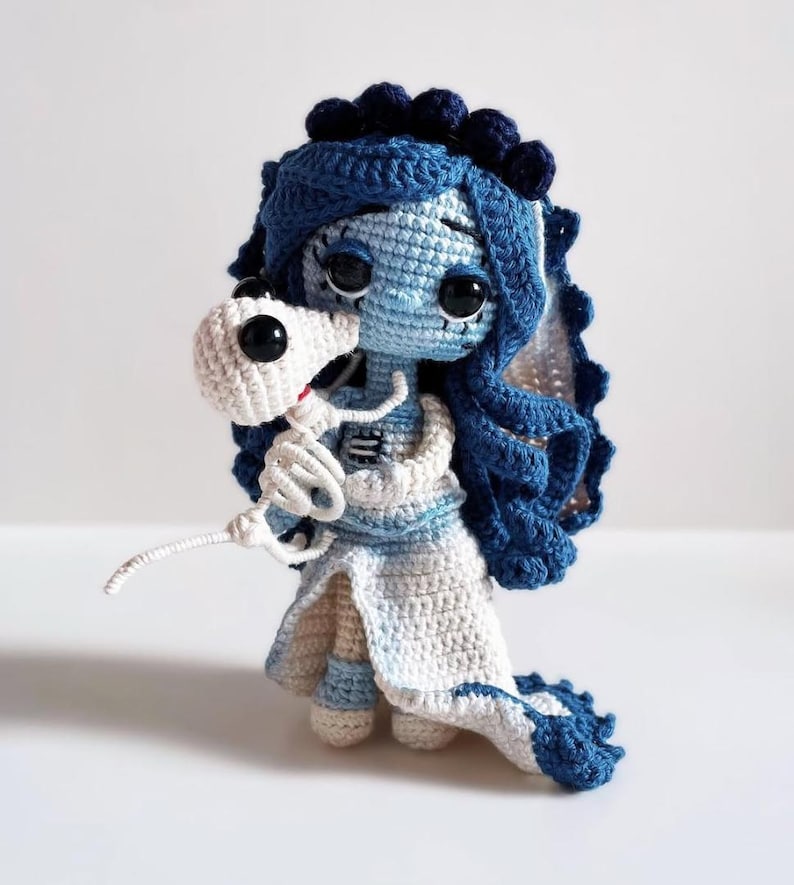 Puede incluir: Mu&ntilde;eca de ganchillo hecha a mano al estilo de La Novia Cad&aacute;ver. La mu&ntilde;eca presenta una combinaci&oacute;n de colores azul y blanco, con un velo y pelo azul, y un vestido blanco. Sostiene un peque&ntilde;o esqueleto blanco. La mu&ntilde;eca tiene ojos grandes y negros.