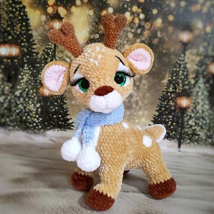 May include: A handmade, plush reindeer with brown antlers, a light brown body with white spots, and a blue scarf with white pom-poms. The reindeer has large green eyes with long eyelashes and pink-lined ears. A festive holiday decoration.