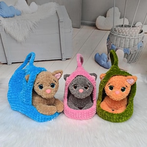 May include: Three plush cat toys in different colors. One is brown in a blue carrier, another is gray in a pink carrier, and the third is orange in a green carrier. Each cat has large blue eyes and a pink nose.