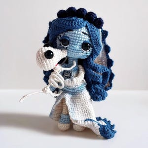 May include: A handmade crochet doll inspired by the Corpse Bride. The doll is predominantly blue and white, with a blue veil and hair, and a white dress. It holds a white skeleton. The doll has large black eyes and is a charming handmade item.