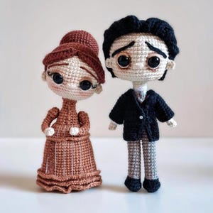 Puede incluir: Dos mu&ntilde;ecas de ganchillo hechas a mano. Una mu&ntilde;eca lleva un vestido marr&oacute;n y tiene el pelo casta&ntilde;o. La otra mu&ntilde;eca lleva un traje negro y tiene el pelo negro. Ambas mu&ntilde;ecas tienen grandes ojos negros y est&aacute;n de pie una al lado de la otra.