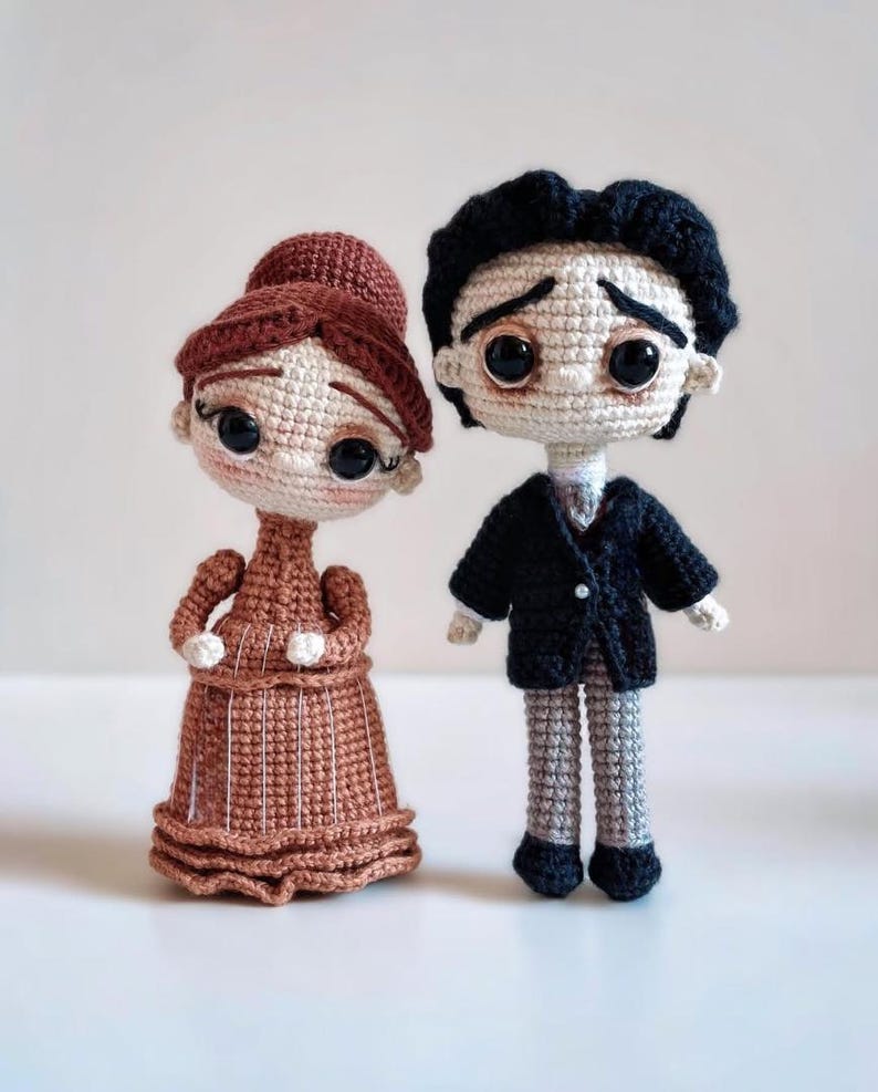 Puede incluir: Dos mu&ntilde;ecas de ganchillo hechas a mano. Una mu&ntilde;eca lleva un vestido marr&oacute;n y tiene el pelo casta&ntilde;o. La otra mu&ntilde;eca lleva un traje negro y tiene el pelo negro. Ambas mu&ntilde;ecas tienen grandes ojos negros y est&aacute;n de pie una al lado de la otra.