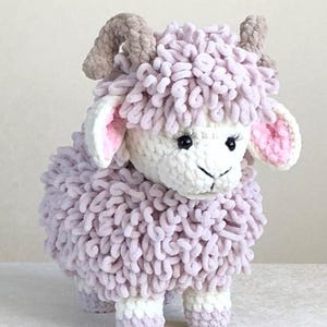 Puede incluir: Un juguete de peluche de oveja hecho a mano con un cuerpo esponjoso de color lavanda y una cara blanca. La oveja tiene cuernos pequeños de color beige, orejas forradas de rosa y ojos negros. El juguete está hecho de hilo suave y texturizado.
