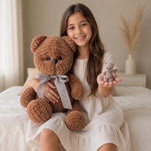 May include: A large, brown, plush, crocheted teddy bear with a grey polka-dot ribbon tied around its neck, accompanied by a tiny, light beige, crocheted miniature teddy bear held in the palm of a hand. Both bears feature dark, round eyes.