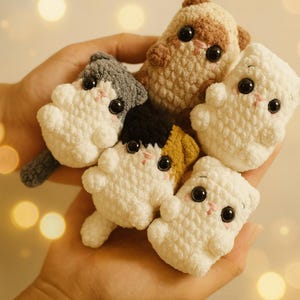 May include: A hand holds a collection of five small, crocheted cat figurines. The cats are primarily white, with accents of gray, brown, black, and yellow. Each cat has large black eyes and pink cheeks, giving them a cute appearance. The background is blurred with bokeh lights.