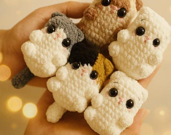 May include: A hand holds a collection of five small, crocheted cat figurines. The cats are primarily white, with accents of gray, brown, black, and yellow. Each cat has large black eyes and pink cheeks, giving them a cute appearance. The background is blurred with bokeh lights.