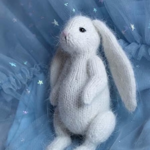 May include: A handmade white knitted bunny toy with long ears, black eyes, and a pink nose. The bunny is sitting on a light blue fabric with star-shaped glitter. The toy is soft and fluffy.