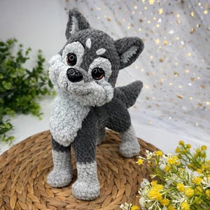 Puede incluir: Un juguete de perro husky tejido a ganchillo, en tonos grises y blancos. El peluche tiene grandes ojos marrones y una nariz negra. El perro está sobre una estera tejida, con vegetación y flores amarillas en el fondo.