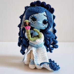 May include: A handcrafted crochet doll with blue hair and a white dress, holding a green and pink snake. The doll has large black eyes and is made with blue, white, and green yarn. The doll's hair is styled in ringlets.