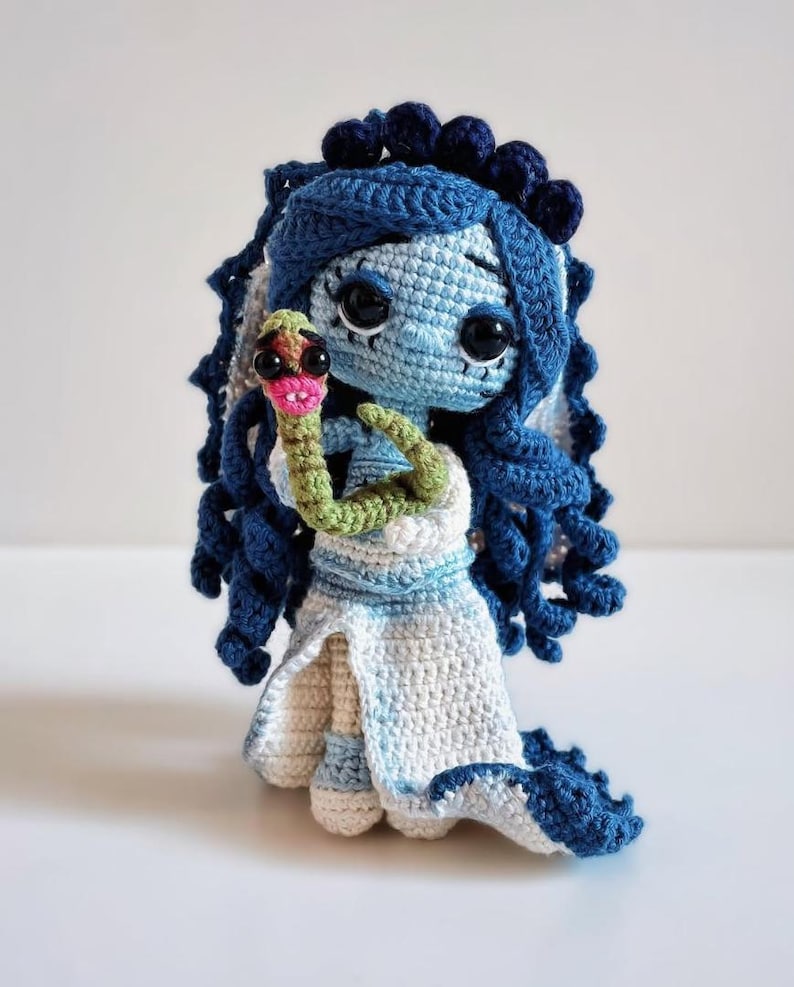 May include: A handcrafted crochet doll with blue hair and a white dress, holding a green and pink snake. The doll has large black eyes and is made with blue, white, and green yarn. The doll's hair is styled in ringlets.
