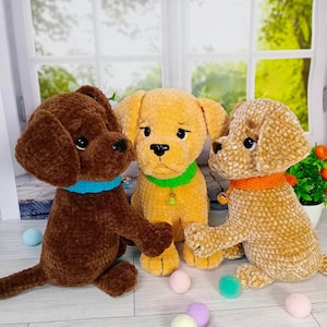 May include: Three plush dog toys in brown, yellow, and tan. Each dog has floppy ears, a black nose, and a colored collar. The brown dog has a blue collar, the yellow dog has a green collar, and the tan dog has an orange collar. Small decorative balls are scattered around the dogs.
