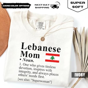 May include: Ivory-colored t-shirt with the text "Lebanese Mom" and a Lebanese flag graphic. The shirt includes a definition of "Mom" and is labeled "Super Soft". A gold necklace is draped across the shirt.