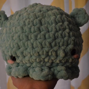 May include: A handmade, light teal crocheted plush toy with small ears, black eyes, and pink cheeks. The plush has a rounded shape and a textured surface, resembling a friendly creature.