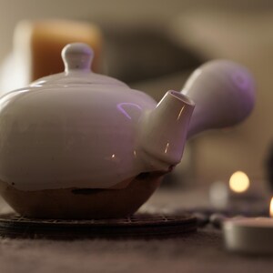 May include: A white ceramic teapot with a rounded body and a spout on the side. The teapot has a brown base and is sitting on a wooden coaster.